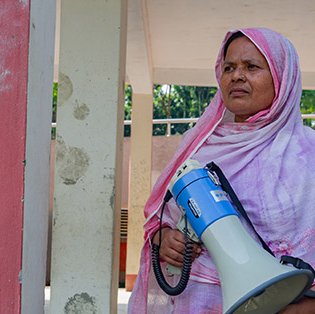 Dulu, a Bangladeshi woman in a pink shawl, stands looking determined while holding a megaphone.