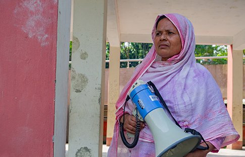 Dulu, a Bangladeshi woman in a pink shawl, stands looking determined while holding a megaphone.