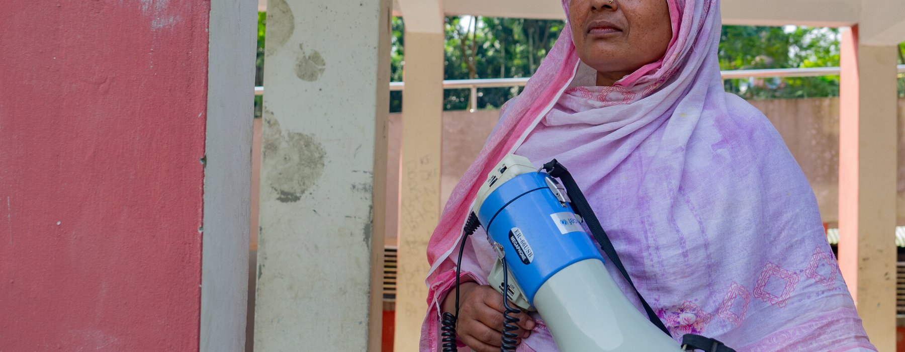 Dulu is wearing a pink hijab and holds a blue megaphone outside a village building.