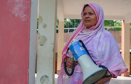 Dulu is wearing a pink hijab and holds a blue megaphone outside a village building.