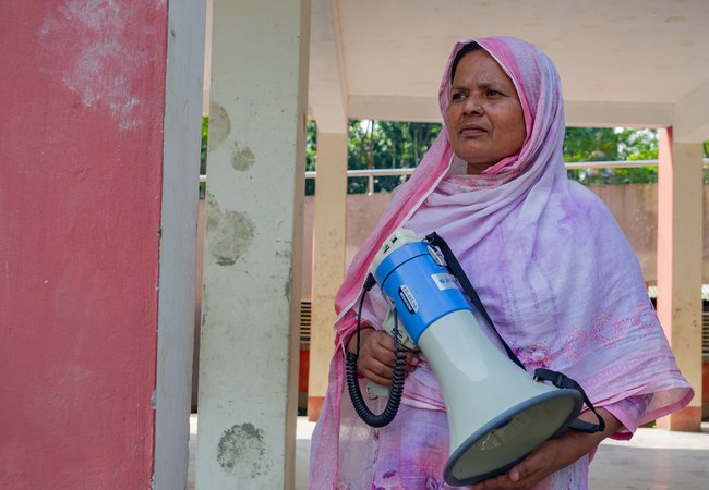 Dulu is wearing a pink hijab and holds a blue megaphone outside a village building.