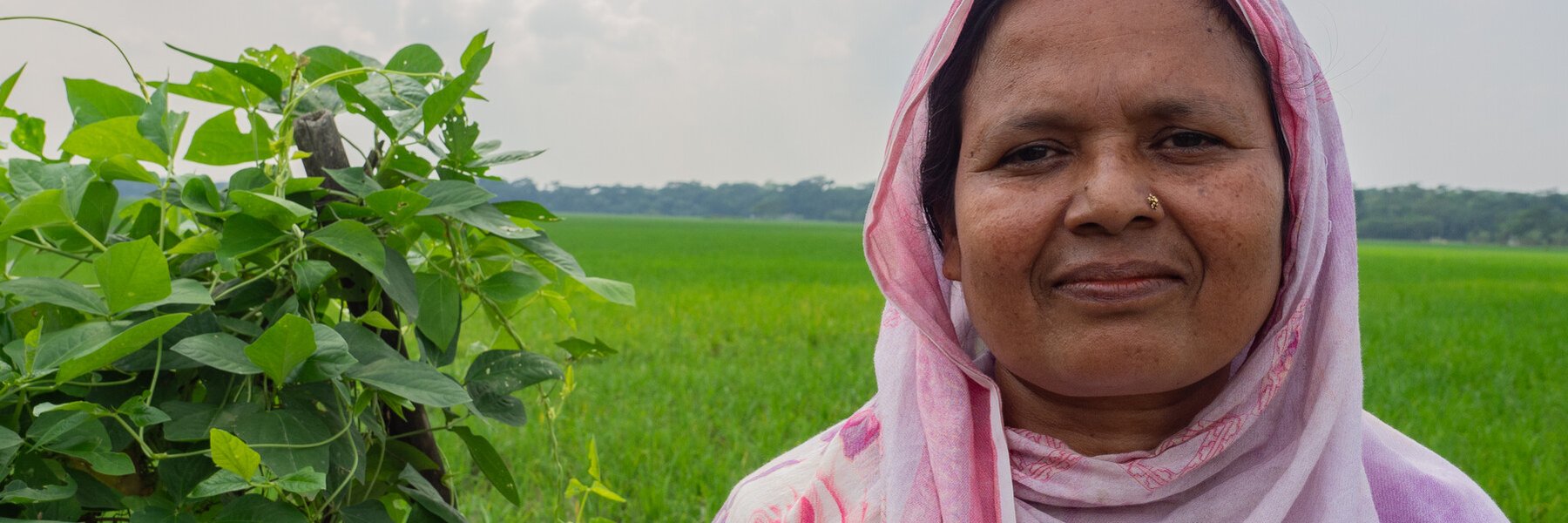 A smiling Bangladeshi woman in a pink shawl stands in the foreground with a luscious green field in the background.