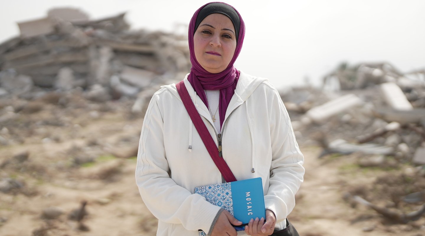 Fidaa Shurrab wearing a purple and black hijab and white coat and carrying a blue notebook with a Middle East kitchen tile pattern on it, stands in front of a destroyed building in Gaza strip while going to work.