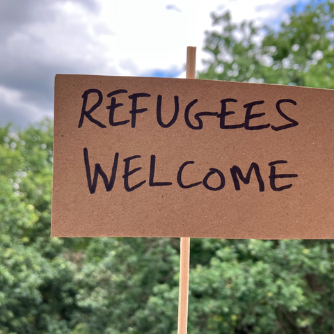 A cardboard placard that says 'refugees welcome' with trees and blue sky in the background