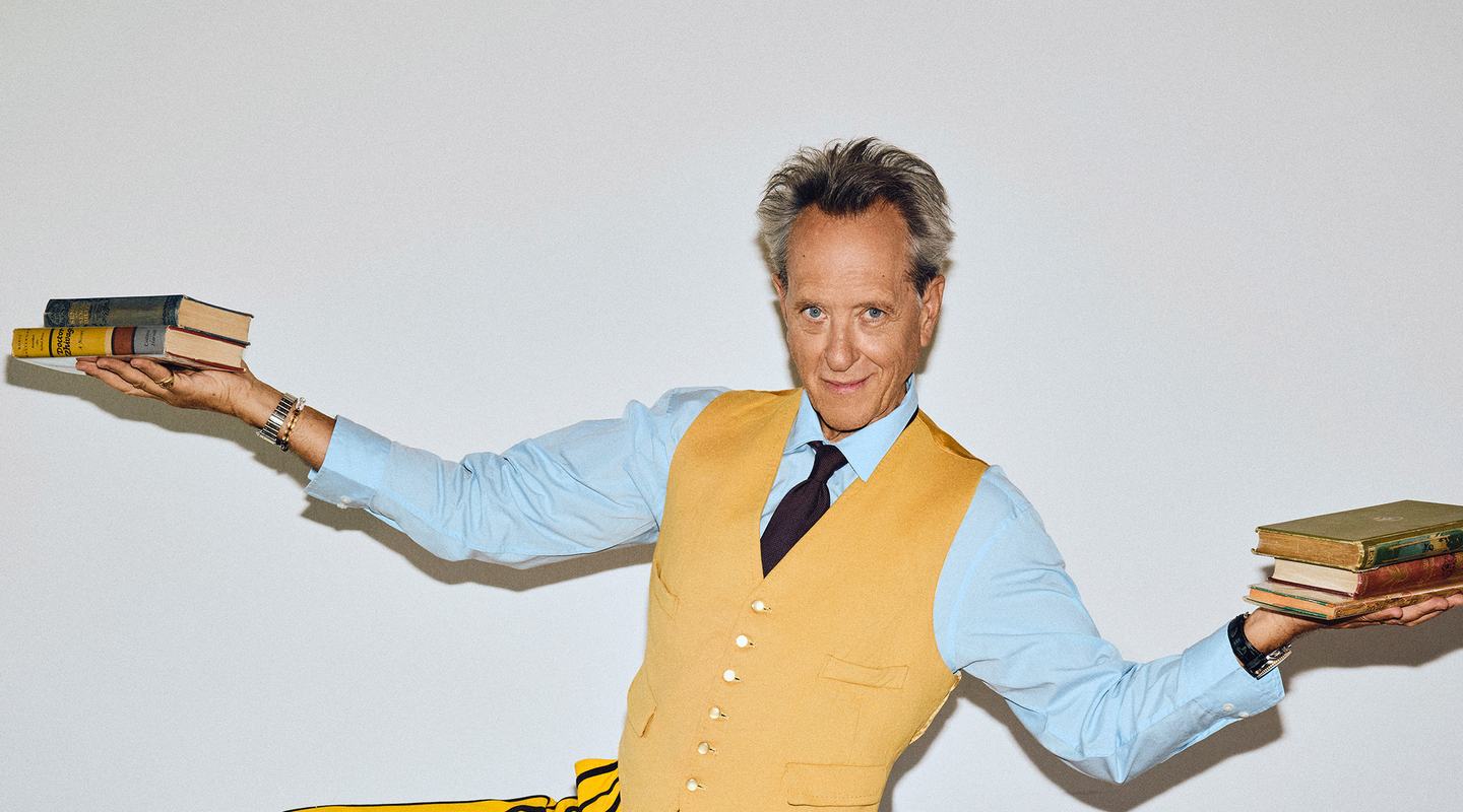 Actor Richard E. Grant, wearing a yellow waistcoat, blue shirt and striped yellow trousers, poses with arms outstretched, balancing stacks of vintage books in each hand.