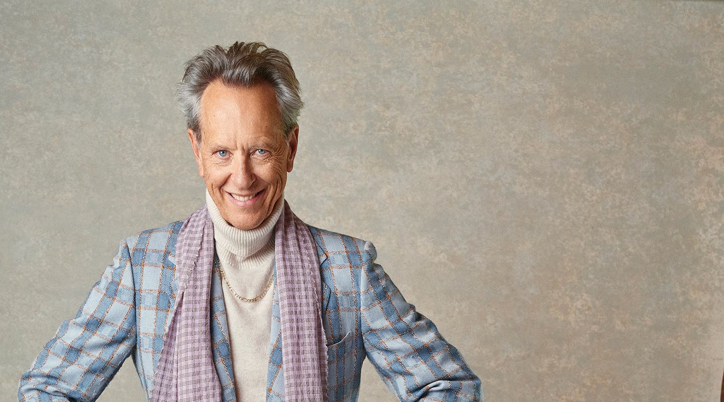 Actor Richard E. Grant stands with his hands on hips in a studio setting, wearing a light blue checked blazer, cream roll-neck sweater, grey trousers, and a lilac scarf. The portrait is set against a neutral textured backdrop.