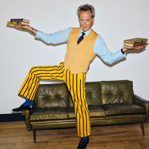 Actor Richard E. Grant, wearing a yellow waistcoat and striped yellow trousers, balances on a green leather sofa while holding stacks of books in both hands.