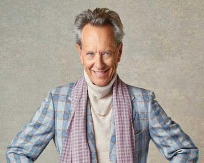 Actor Richard E. Grant stands with his hands on hips in a studio setting, wearing a light blue checked blazer, cream roll-neck sweater, grey trousers, and a lilac scarf. The portrait is set against a neutral textured backdrop.