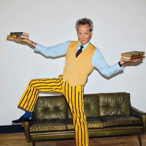 Actor Richard E. Grant, wearing a yellow waistcoat and striped yellow trousers, balances on a green leather sofa while holding stacks of books in both hands.