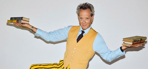 Actor Richard E. Grant, wearing a yellow waistcoat and striped yellow trousers, balances on a green leather sofa while holding stacks of books in both hands.