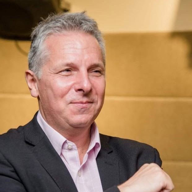 A person wearing a suit jacket and light-coloured shirt sits indoors with one arm raised across their chest.