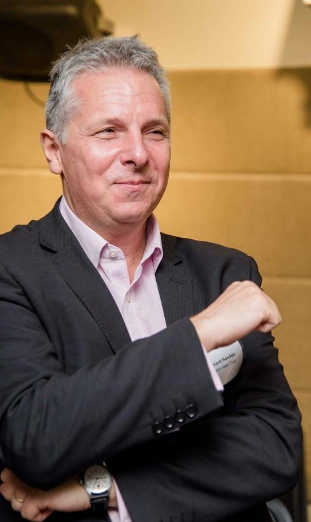A person wearing a suit jacket and light-coloured shirt sits indoors with one arm raised across their chest.
