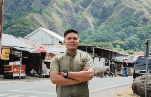 A man with short dark hair and a green shirt stands with his arms crossed with green hills in the background.