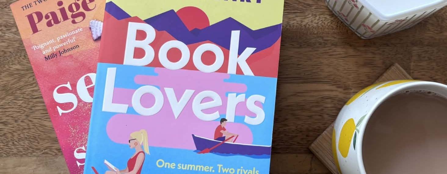 Two romance novels, 'Book Lovers' by Emily Henry and 'Seven Summers' by Paige Toon, beside a mug of tea and a candle on a wooden table.