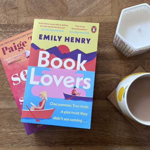 Two romance novels, 'Book Lovers' by Emily Henry and 'Seven Summers' by Paige Toon, beside a mug of tea and a candle on a wooden table.