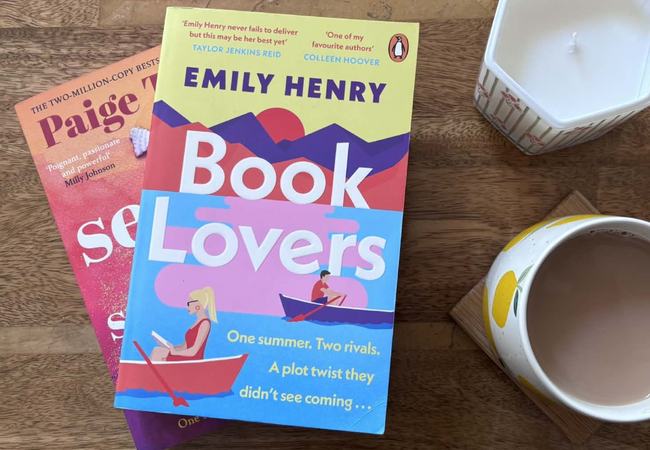 Two romance novels, 'Book Lovers' by Emily Henry and 'Seven Summers' by Paige Toon, beside a mug of tea and a candle on a wooden table.