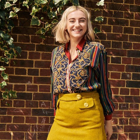 A woman stood smiling in front of a brick wall with leaf's featuring. The woman stands in a yellow skirt with a pocket and a button on it, she also wears a multi-coloured patterned blouse.