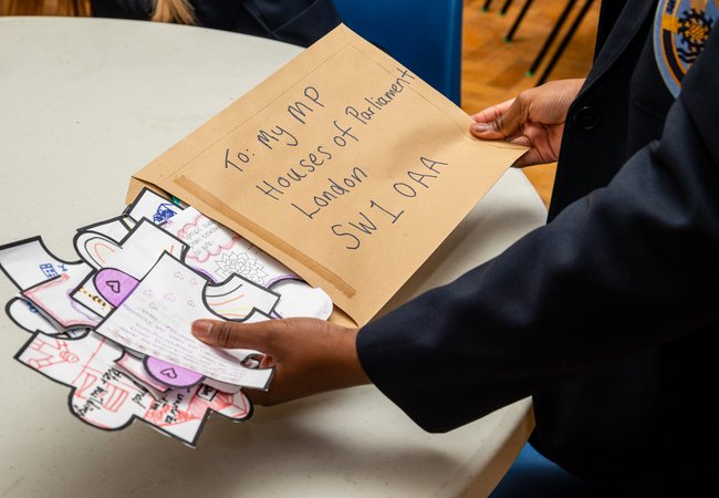 A brown envelope with: To: My Mp, Houses of Parliament, London, SW1 OAA is backed with drawings and writing on paper in the shape of giant puzzle pieces by a child.