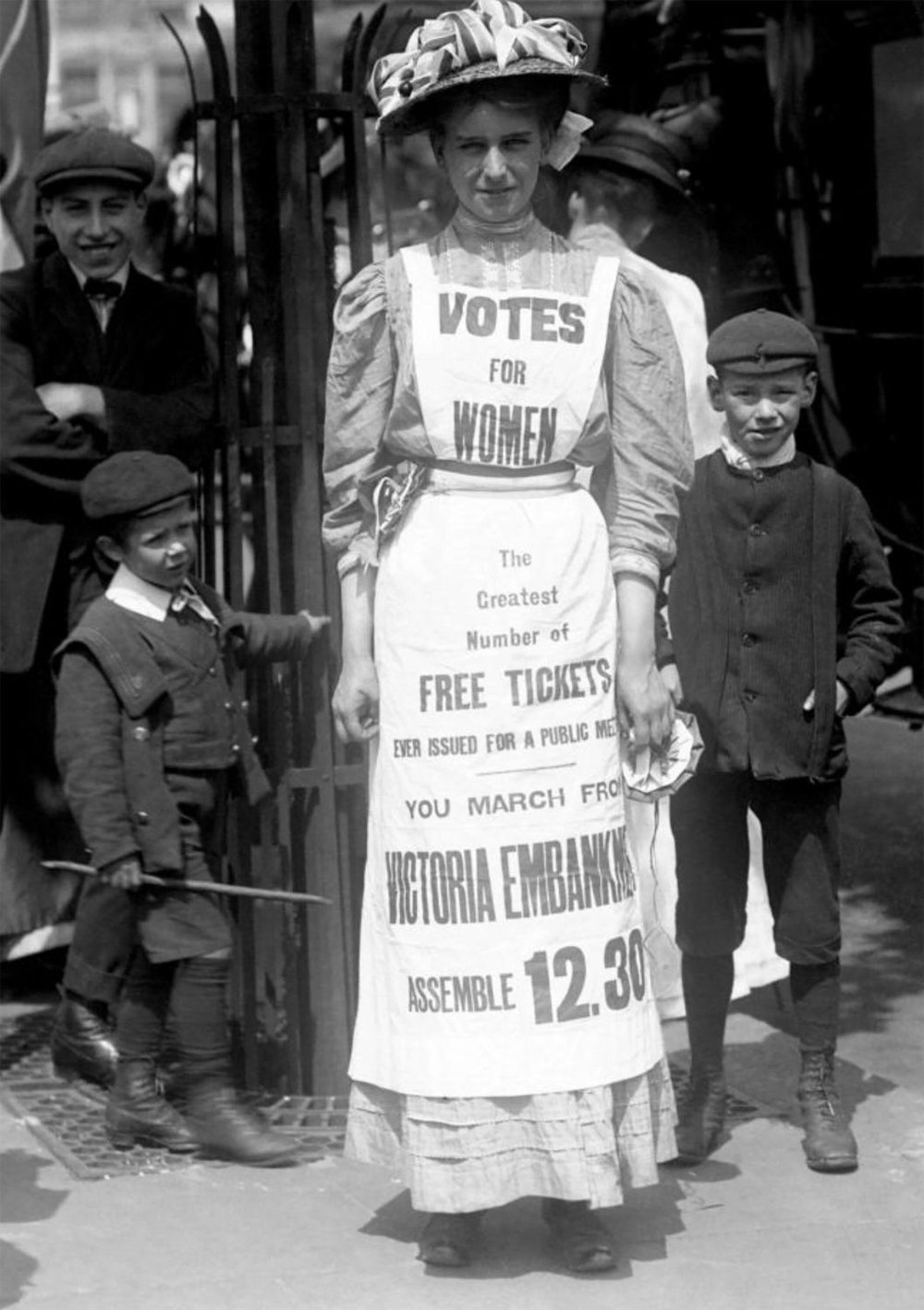 A black-and-white photo of Vera Wentworth, a suffragette. She wears a hat, a long dress, and a long apron emblazoned with text that publicises a protest march.