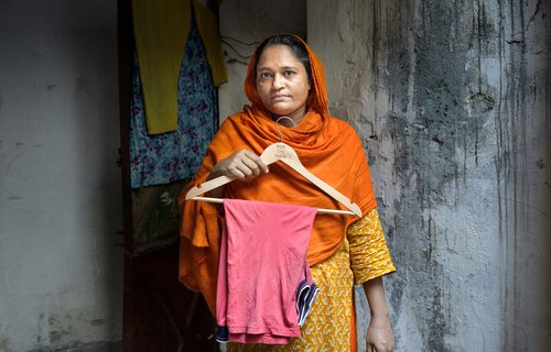 A woman in an orange head scarf holds a clothes hanger containing a garment she has made.