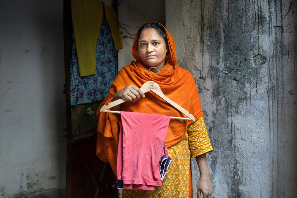 A woman in an orange head scarf holds a clothes hanger containing a garment she has made.