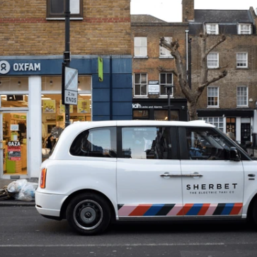 A white electric taxi with “SHERBET” branding is parked on a street in front of an Oxfam store in an urban area.