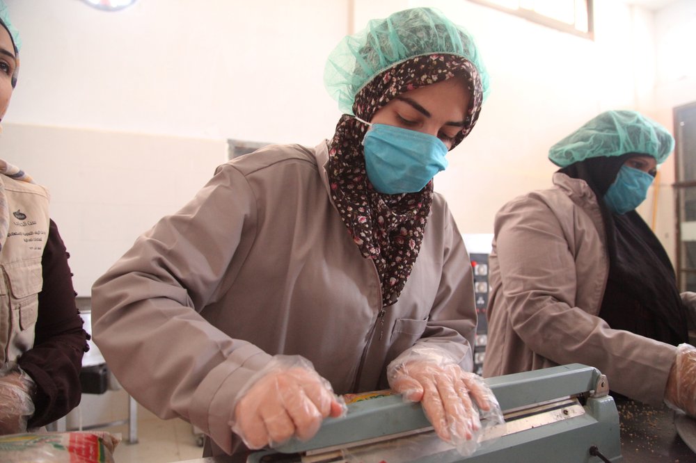 Shatha wears a blue hair net and mask and a flowery headscarf and uses a packaging guillotine to package food