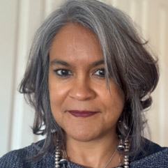 A woman with shoulder-length grey hair, wearing a dark top and beaded necklace, facing the camera indoors.