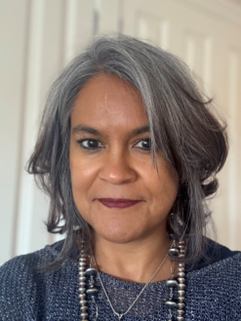 A woman with shoulder-length grey hair, wearing a dark top and beaded necklace, facing the camera indoors.