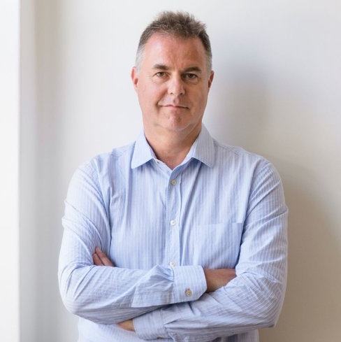 A man wearing a striped light blue dress shirt and jeans looks into the camera, with his arms folded, against a light coloured wall.