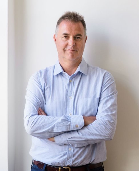 A man wearing a striped light blue dress shirt and jeans looks into the camera, with his arms folded, against a light coloured wall.