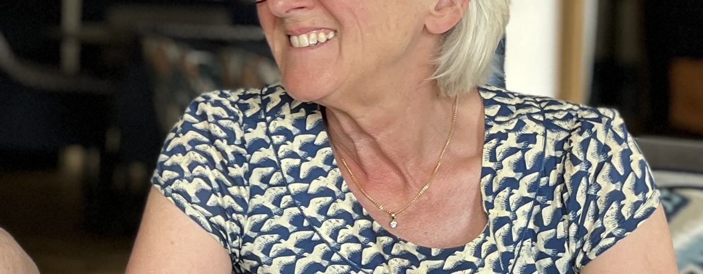 Smiling person with short grey hair and glasses seated at a table, looking to the side, wearing a patterned blue top.