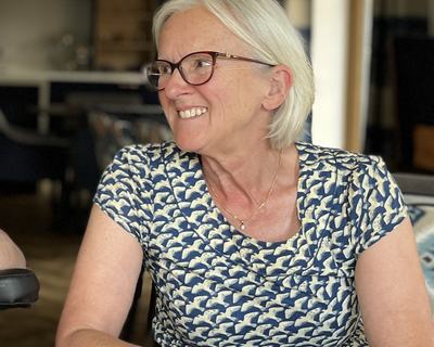 Smiling person with short grey hair and glasses seated at a table, looking to the side, wearing a patterned blue top.