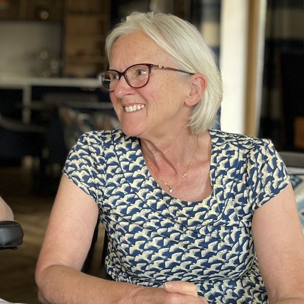 Smiling person with short grey hair and glasses seated at a table, looking to the side, wearing a patterned blue top.