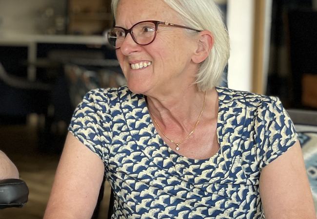Smiling person with short grey hair and glasses seated at a table, looking to the side, wearing a patterned blue top.