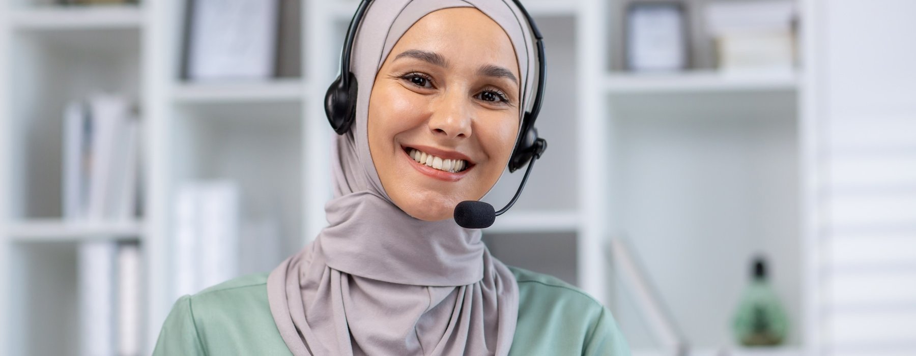 A smiling woman looks straight into the camera. She is wearing a light grey headscarf and a headset, sitting in front of a laptop.