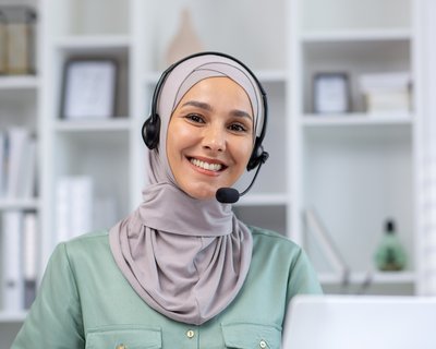 A smiling woman looks straight into the camera. She is wearing a light grey headscarf and a headset, sitting in front of a laptop.