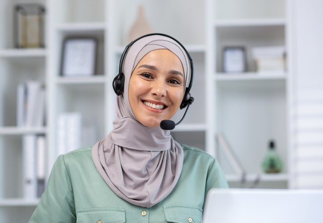 A smiling woman looks straight into the camera. She is wearing a light grey headscarf and a headset, sitting in front of a laptop.