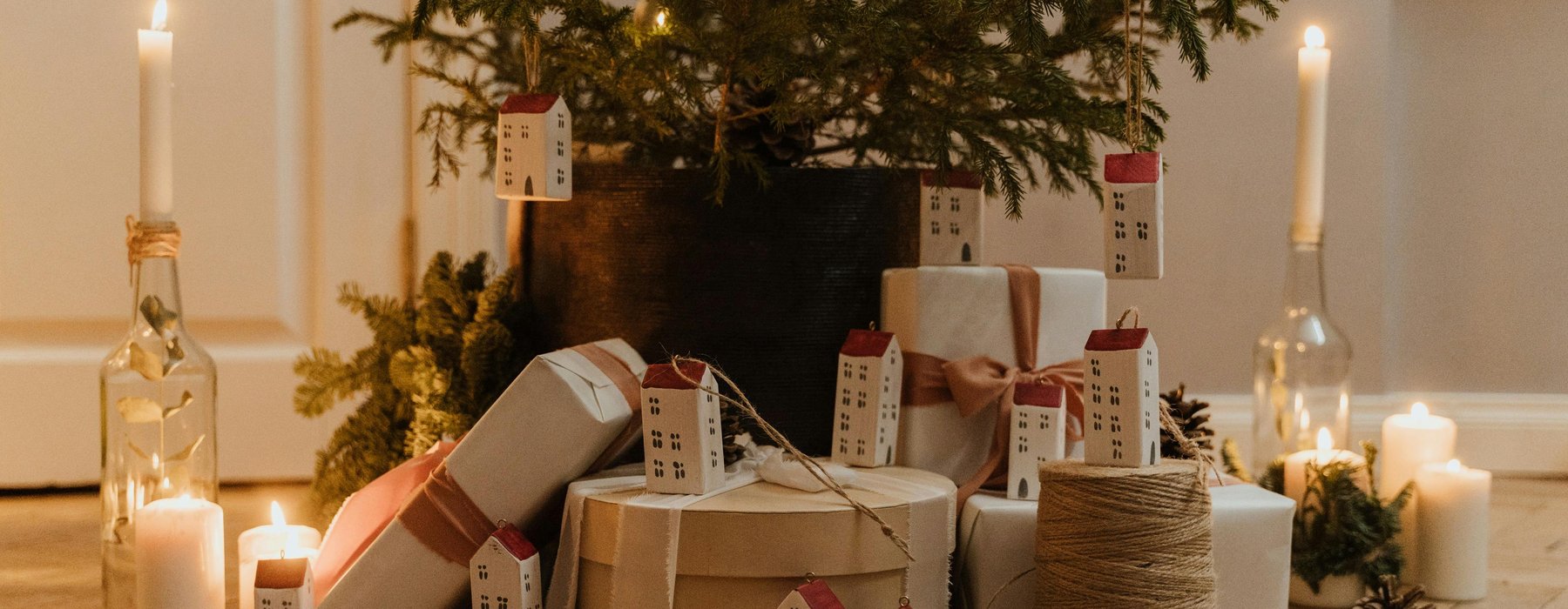 Christmas tree with yellow lights, place in a wooden floor with presents wrapped in brown paper. The tree has minimal decorations apart from the lights and long white house decorations hanging off. There is also candles surrounding the tree.