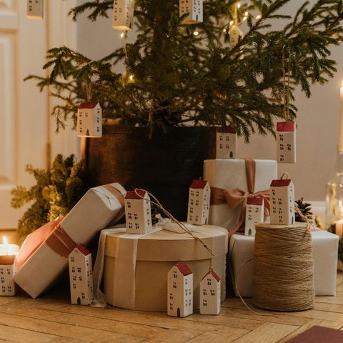 Christmas tree with yellow lights, place in a wooden floor with presents wrapped in brown paper. The tree has minimal decorations apart from the lights and long white house decorations hanging off. There is also candles surrounding the tree.