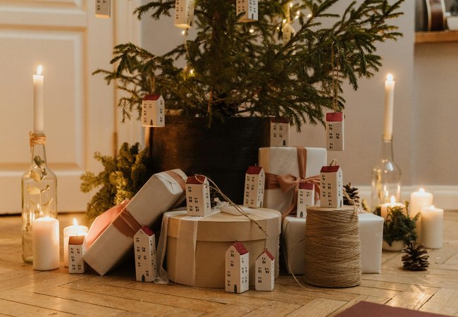 Christmas tree with yellow lights, place in a wooden floor with presents wrapped in brown paper. The tree has minimal decorations apart from the lights and long white house decorations hanging off. There is also candles surrounding the tree.