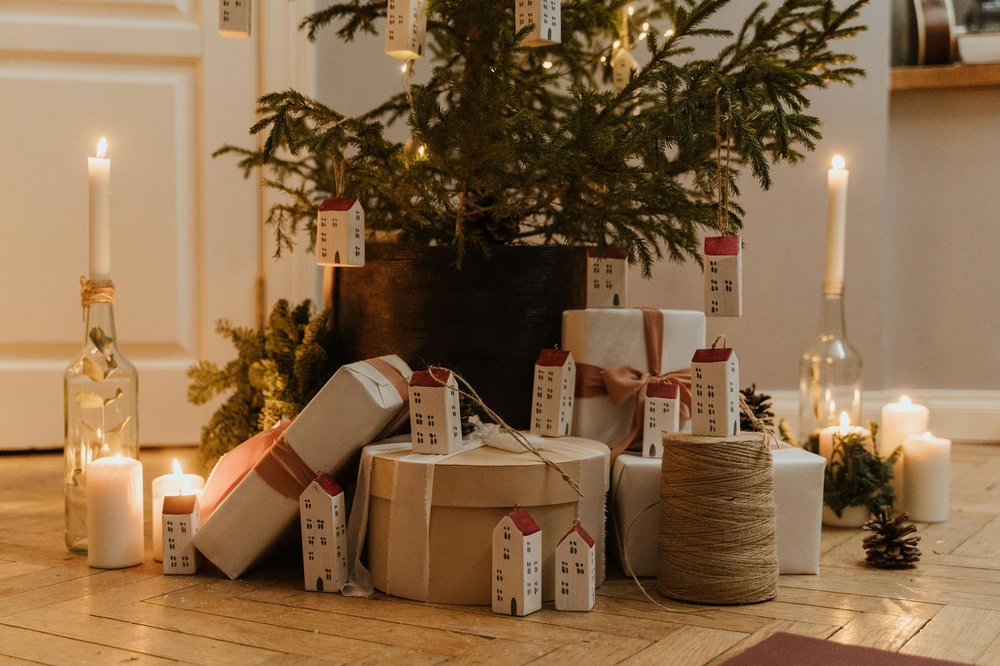 Christmas tree with yellow lights, place in a wooden floor with presents wrapped in brown paper. The tree has minimal decorations apart from the lights and long white house decorations hanging off. There is also candles surrounding the tree.