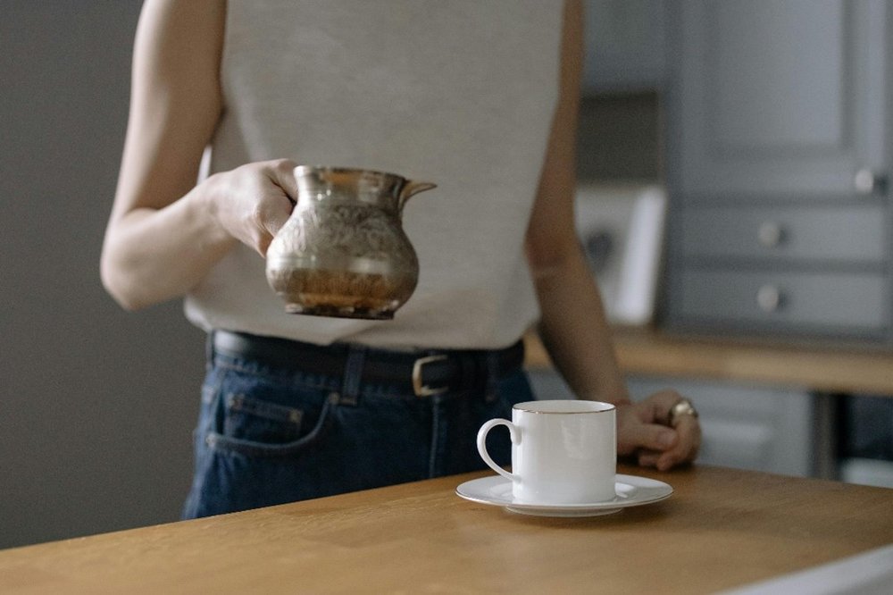 Person in a kitchen near the table holding a antique teapot and with a white china tea cup and sauce placed on the wooden surface.