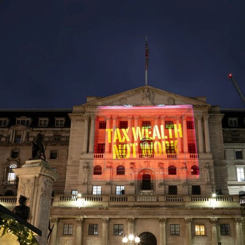 The Bank of England made of stone brick featuring stone pillars and a balcony with a flagpole on the top flying the flag of the UK. There is a red projection on the front of the building with yellow text that says "Tax wealth not work"