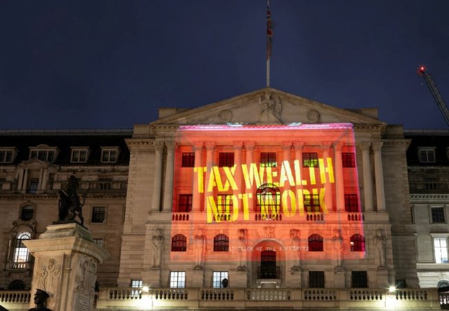 The Bank of England made of stone brick featuring stone pillars and a balcony with a flagpole on the top flying the flag of the UK. There is a red projection on the front of the building with yellow text that says "Tax wealth not work"
