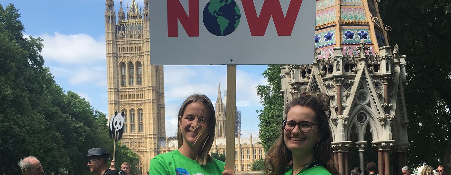 Two Oxfam staff members stand outside the Houses of Parliament holding up a placard saying 'The Time is Now'.