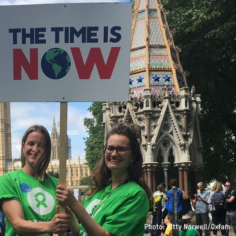 Two Oxfam staff members stand outside the Houses of Parliament holding up a placard saying 'The Time is Now'.