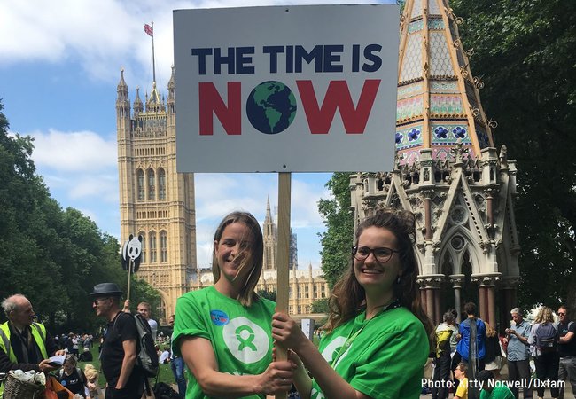 Two Oxfam staff members stand outside the Houses of Parliament holding up a placard saying 'The Time is Now'.