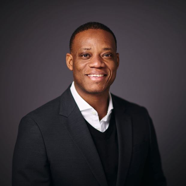 A man wearing a white shirt, black jumper and black suit jacket smiles into the camera. The background is plain dark grey.