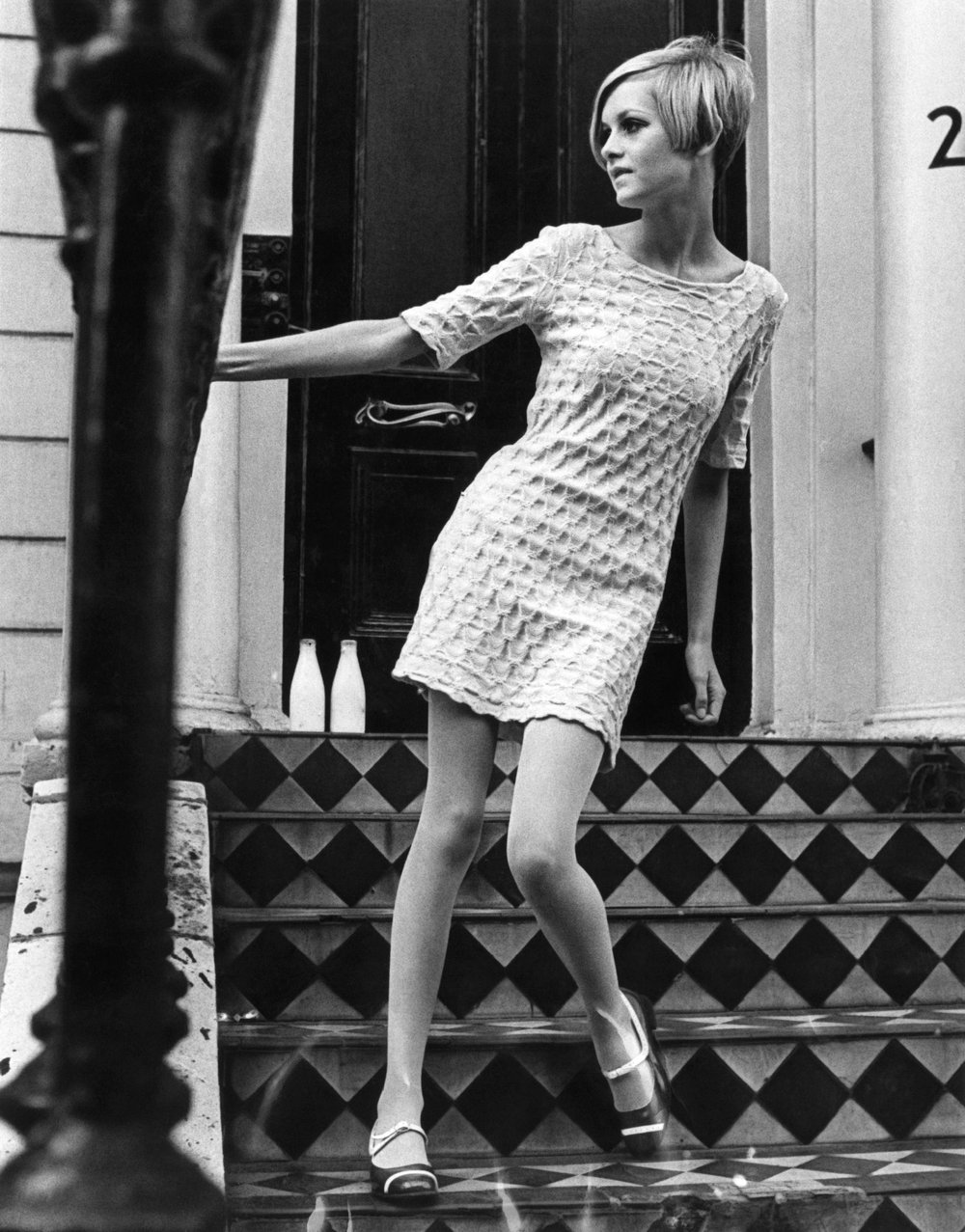 A black-and-white photo of model Twiggy, standing on steps that have a geometric pattern. She has cropped hair and wears a short dress and Mary Jane shoes.